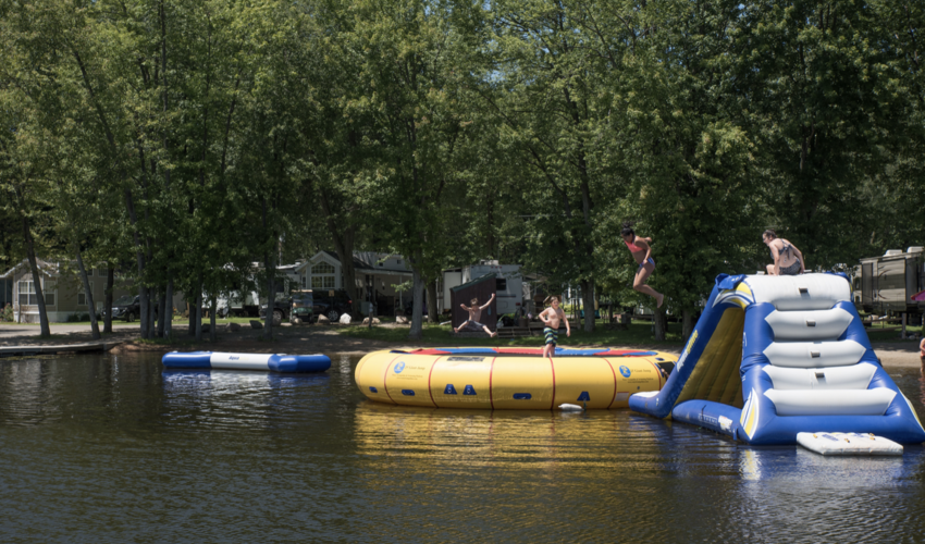 Four children jumping on a collection of inflatable floating platforms in a lake surrounded by trees