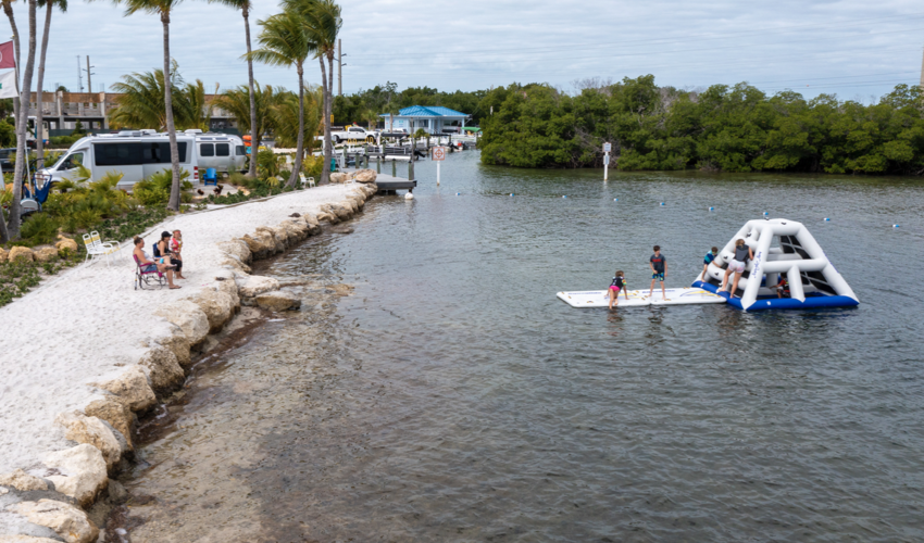 Children playing in an inflatable structure on a lake surrounded by palm trees and a campground.