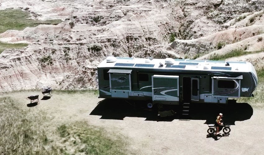A fifth wheel trailer parked in a badlands landscape, with a bicyclist riding past it, as viewed from above