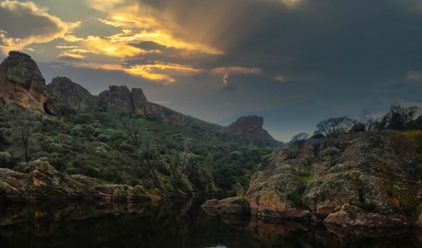 Tall rock formations and greenery rise out of a dark lake, with a sunset partially obscured by clouds above it.