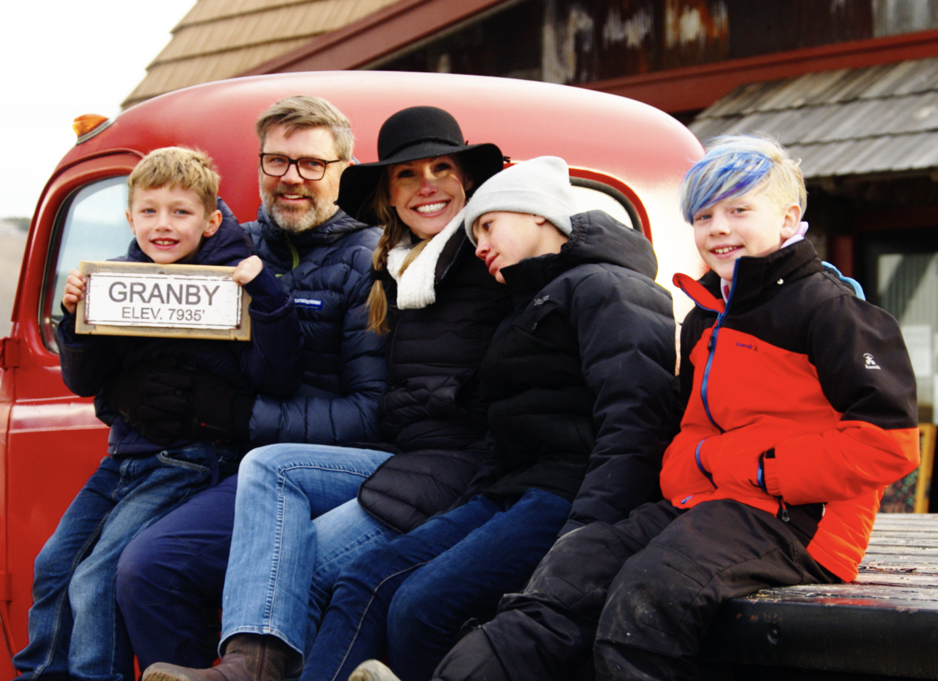 A family of five sits on a truck bed holding up a sign that reads “Granby, Elevation 7,935 feet”