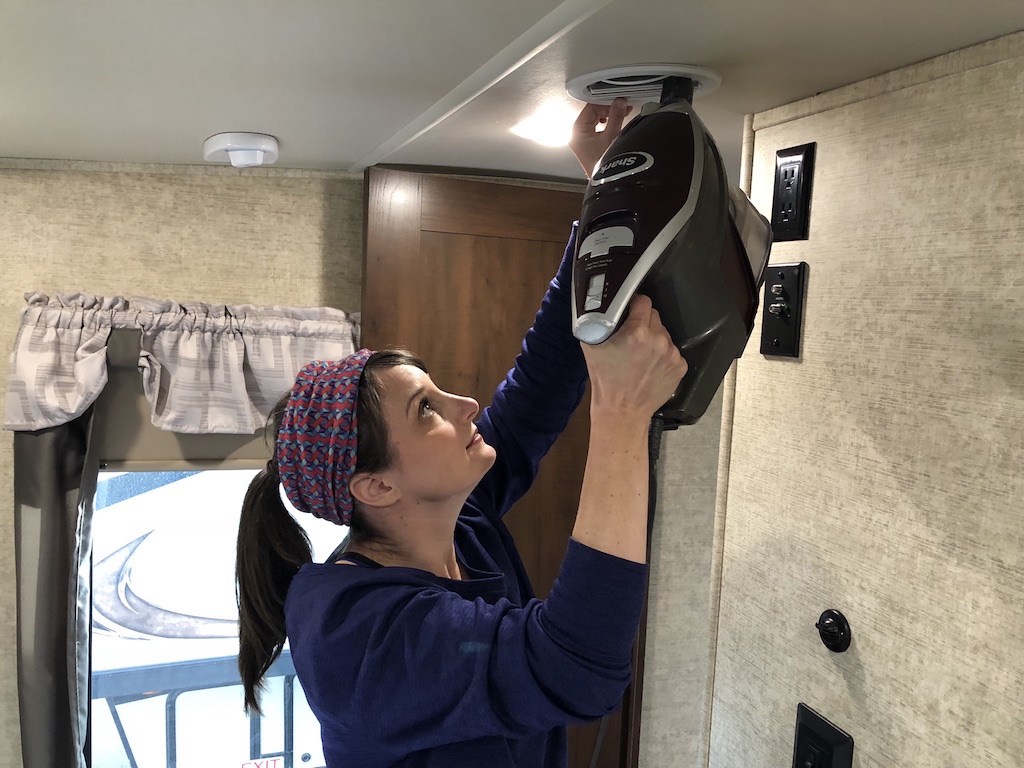 Person using a vacuum attachment to clean dust and debris from the ceiling vents of an RV interior.