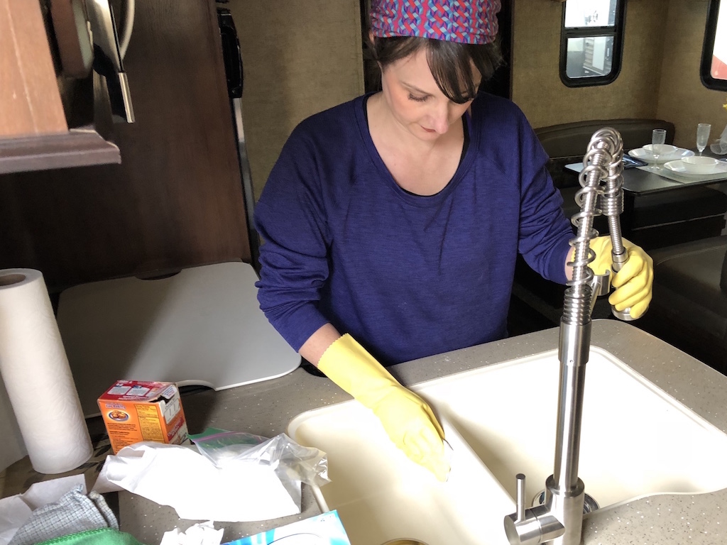 Person wearing yellow gloves cleaning an RV kitchen sink using a sponge and biodegradable cleaner.