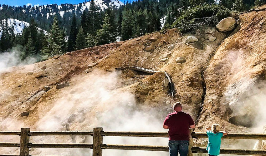 A man and a child stand with their backs against the camera, looking at a geological feature with rising steam and snow-covered mountains in the background